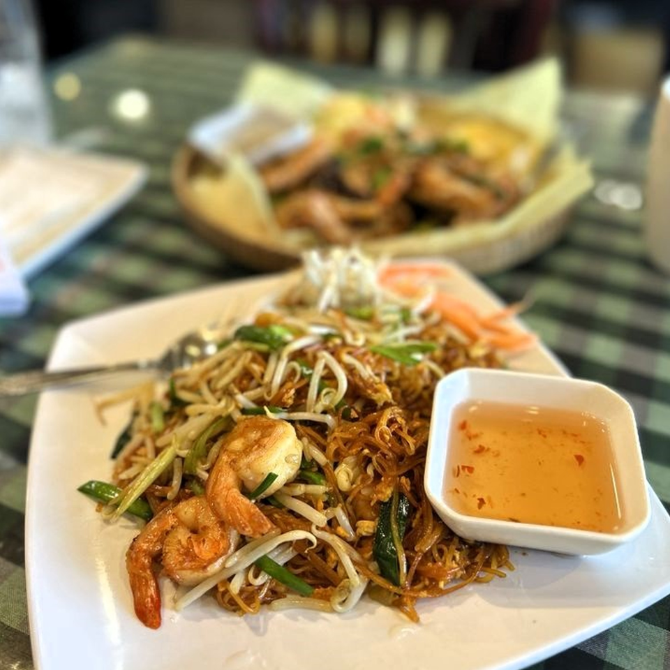 Cambodian noodle dish with shrimp, bean sprouts, and a side of dipping sauce on a white plate.